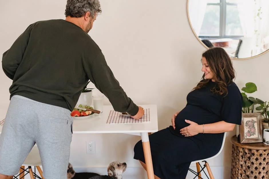Side view of smiling pregnant female in casual clothes sitting at table while husband putting plate with food in kitchen in cozy apartment